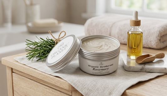A silver tin of Organic Deodorant Rosemary sits on a linen surface beside a small dropper bottle of oil, a wooden spoon, and a sprig of rosemary in a softly lit bathroom-like setting.